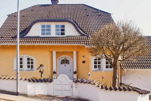 Gelbes Haus mit Satteldach und weißen Fenstern, umgeben von einem niedrigen Zaun.